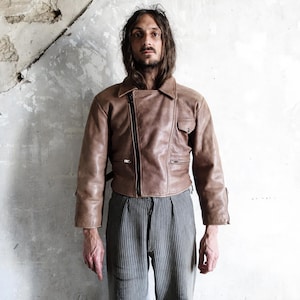 Puede incluir: Chaqueta de cuero marrón con cierre de cremallera asimétrico y un pequeño bolsillo en el pecho. La chaqueta tiene un estilo motero clásico con cuello de solapa y mangas largas. El modelo lleva la chaqueta con pantalones a rayas.