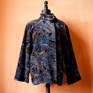 Puede incluir: Una chaqueta de terciopelo azul con estampado paisley. La chaqueta tiene un cuello mandarín y cierre de botón.
