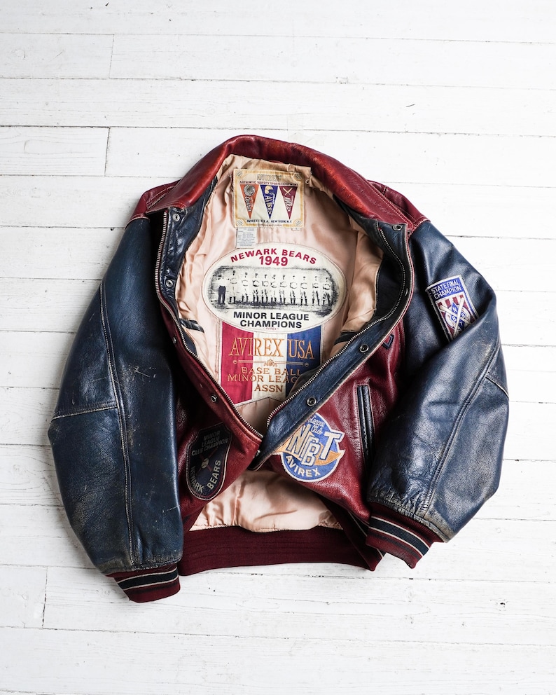 May include: A red and blue leather varsity jacket featuring a beige interior lining with printed graphics including Newark Bears 1949 Minor League Champions, Avirex USA, and various baseball-themed patches on the exterior and interior panels.