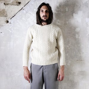 May include: A cream-colored cable knit sweater with a crew neck. The sweater has a textured pattern and long sleeves. The person in the image is wearing gray pants. The background is a distressed, textured wall.