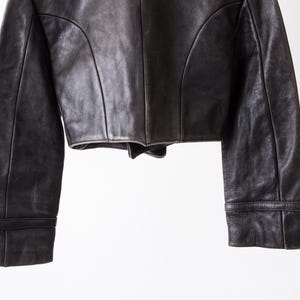 K&ouml;nnte beinhalten: Eine dunkelbraune Lederjacke mit kurzem Schnitt, langen &Auml;rmeln mit strukturierten B&uuml;ndchen und markanten vertikalen Nahtdetails auf den Vorderteilen. Das Kleidungsst&uuml;ck weist eine glatte, matte Textur und eine klare Verarbeitung auf.