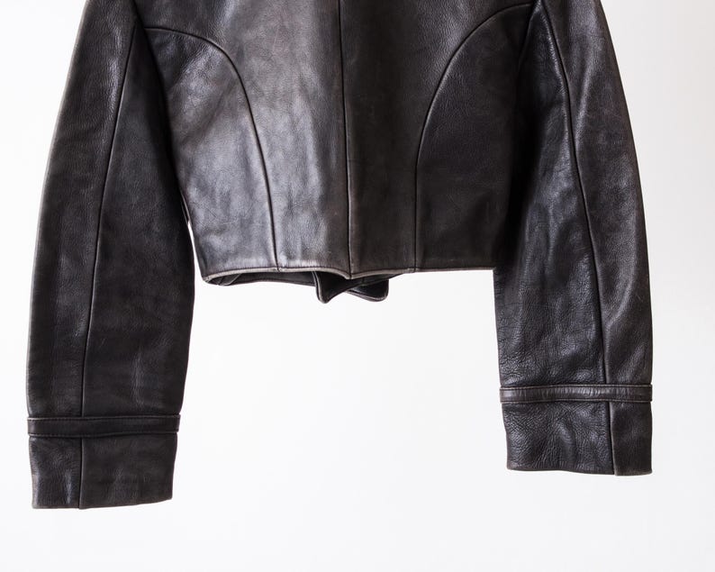 K&ouml;nnte beinhalten: Eine dunkelbraune Lederjacke mit kurzem Schnitt, langen &Auml;rmeln mit strukturierten B&uuml;ndchen und markanten vertikalen Nahtdetails auf den Vorderteilen. Das Kleidungsst&uuml;ck weist eine glatte, matte Textur und eine klare Verarbeitung auf.