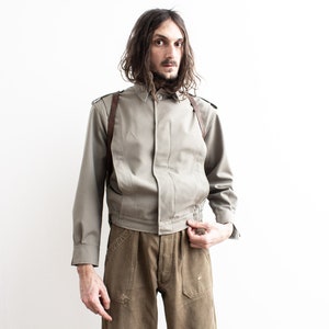 Pu&ograve; includere: Giacca verde oliva chiaro con maniche lunghe e colletto button-down. La giacca ha cinghie in pelle marrone ed &egrave; abbinata a pantaloni marroni. Il modello &egrave; in piedi davanti a uno sfondo bianco.