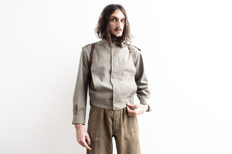 Pu&ograve; includere: Giacca verde oliva chiaro con maniche lunghe e colletto button-down. La giacca ha cinghie in pelle marrone ed &egrave; abbinata a pantaloni marroni. Il modello &egrave; in piedi davanti a uno sfondo bianco.