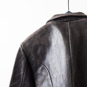 K&ouml;nnte beinhalten: Eine dunkelbraune Lederjacke mit umgelegtem Kragen, sichtbaren Nahtdetails an Schultern und R&uuml;cken sowie einer strukturierten, abgenutzten Oberfl&auml;che. Das Kleidungsst&uuml;ck h&auml;ngt an einem Kleiderb&uuml;gel und zeigt die Konstruktion des oberen R&uuml;ckens.
