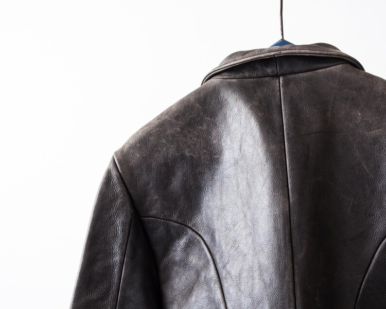 K&ouml;nnte beinhalten: Eine dunkelbraune Lederjacke mit umgelegtem Kragen, sichtbaren Nahtdetails an Schultern und R&uuml;cken sowie einer strukturierten, abgenutzten Oberfl&auml;che. Das Kleidungsst&uuml;ck h&auml;ngt an einem Kleiderb&uuml;gel und zeigt die Konstruktion des oberen R&uuml;ckens.