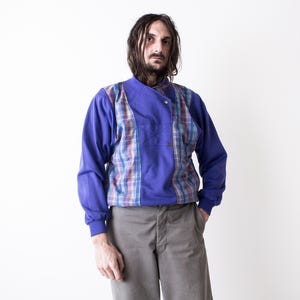 May include: A blue long-sleeve shirt with a plaid panel on the chest. The shirt has a buttoned collar and cuffs. The person is wearing gray pants.