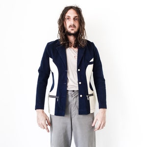 Puede incluir: Un hombre con un blazer azul y blanco con estampado, mangas largas y cierre de bot&oacute;n. Lleva pantalones grises y tiene el pelo largo y casta&ntilde;o.
