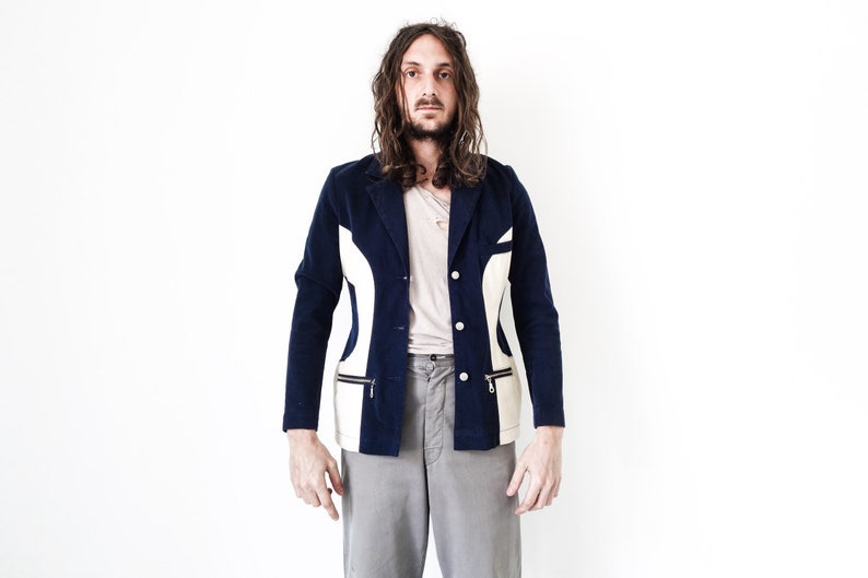 Puede incluir: Un hombre con un blazer azul y blanco con estampado, mangas largas y cierre de bot&oacute;n. Lleva pantalones grises y tiene el pelo largo y casta&ntilde;o.