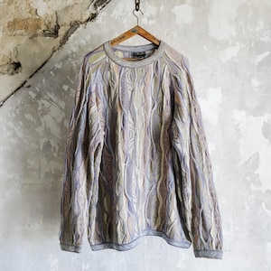 Könnte beinhalten: Ein langärmliger Pullover mit einem einzigartigen, strukturierten Muster in Blau-, Beige- und Grautönen hängt an einem Holzbügel. Der Pullover hat einen Rundhalsausschnitt und gerippte Bündchen und Saum. Der Hintergrund ist eine verwitterte, strukturierte Wand.