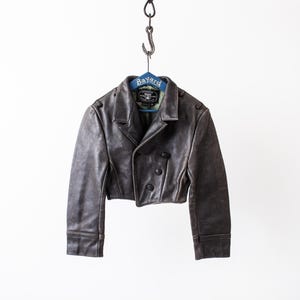 K&ouml;nnte beinhalten: Eine kurze, dunkelbraune Lederjacke mit breitem Kragen, langen &Auml;rmeln und einer zweireihigen Vorderseite mit drei sichtbaren schwarzen Kn&ouml;pfen. Die Jacke h&auml;ngt an einem blauen Kleiderb&uuml;gel mit der Aufschrift Bayard an einem Metallhaken.