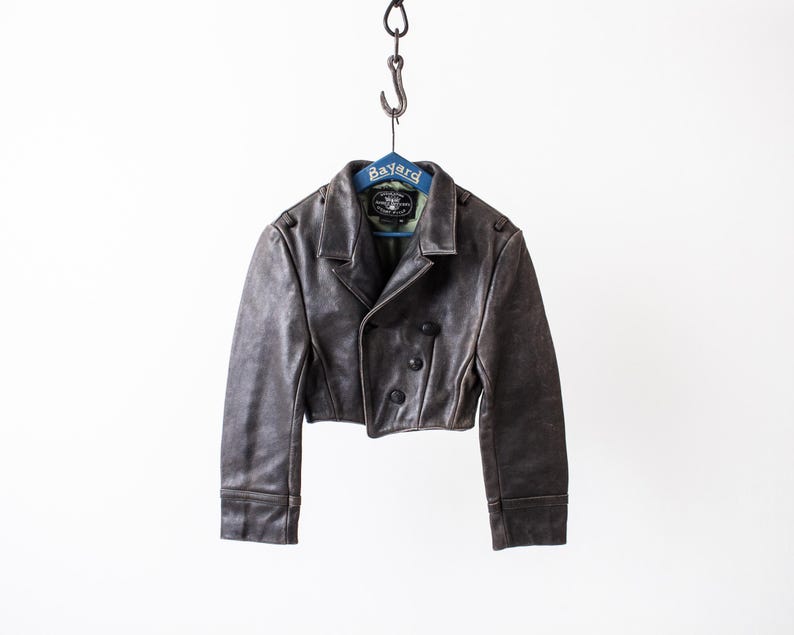 K&ouml;nnte beinhalten: Eine kurze, dunkelbraune Lederjacke mit breitem Kragen, langen &Auml;rmeln und einer zweireihigen Vorderseite mit drei sichtbaren schwarzen Kn&ouml;pfen. Die Jacke h&auml;ngt an einem blauen Kleiderb&uuml;gel mit der Aufschrift Bayard an einem Metallhaken.