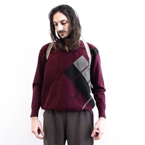 Pode incluir: Uma camisola bordô com um design geométrico em cinza e preto. A camisola tem mangas compridas e decote em V. A pessoa está usando calças marrons e uma alça de cor clara.