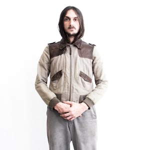 May include: A man wearing a beige and brown bomber jacket with a zipper closure. The jacket has two pockets on the front and a ribbed collar and cuffs.