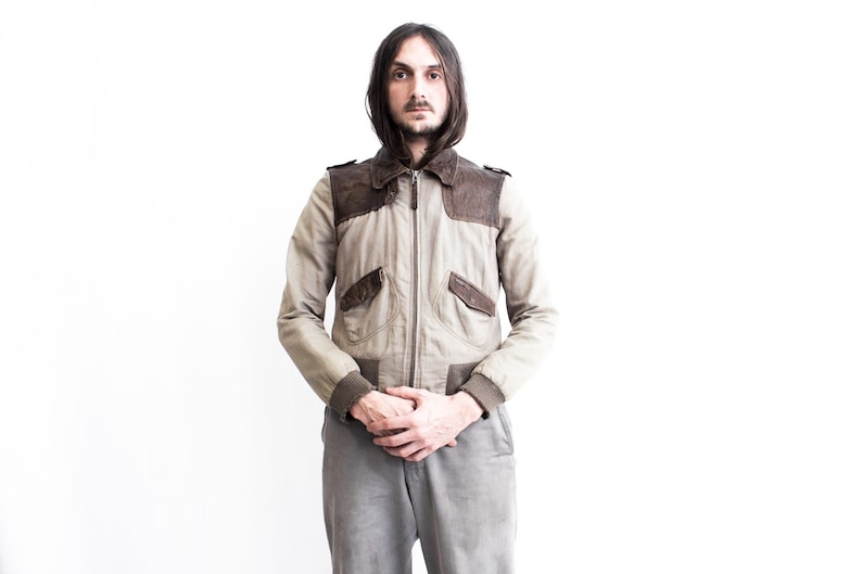 May include: A man wearing a beige and brown bomber jacket with a zipper closure. The jacket has two pockets on the front and a ribbed collar and cuffs.