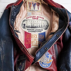May include: A red and blue leather varsity jacket featuring a beige interior lining with a Newark Bears 1949 Minor League Champions patch, an Avirex USA logo, and various baseball-themed embroidered patches on the exterior and interior panels.