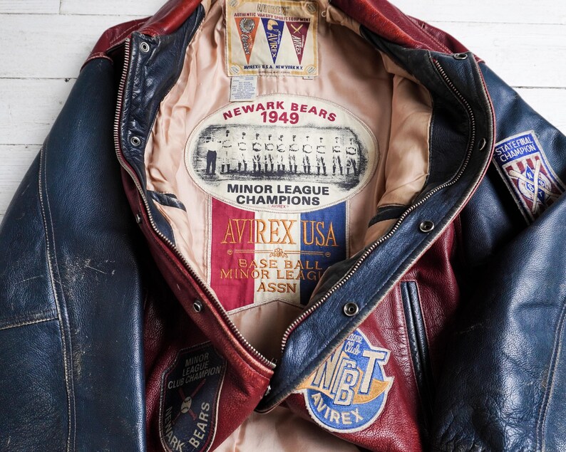 May include: A red and blue leather varsity jacket featuring a beige interior lining with a Newark Bears 1949 Minor League Champions patch, an Avirex USA logo, and various baseball-themed embroidered patches on the exterior and interior panels.