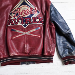 May include: A maroon leather varsity jacket with navy blue sleeves. The back features a large embroidered patch with the text Newark Bears and Avirex, along with a graphic of two crossed baseball bats and the text Minor League Champions.