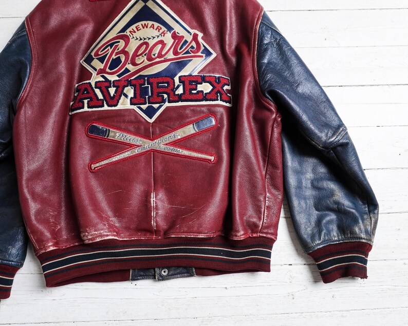 May include: A maroon leather varsity jacket with navy blue sleeves. The back features a large embroidered patch with the text Newark Bears and Avirex, along with a graphic of two crossed baseball bats and the text Minor League Champions.