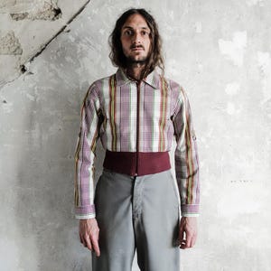 May include: A cropped, collared jacket with a plaid pattern in red, yellow, and white, featuring a burgundy waist band and a zipper closure. The jacket is paired with gray trousers.