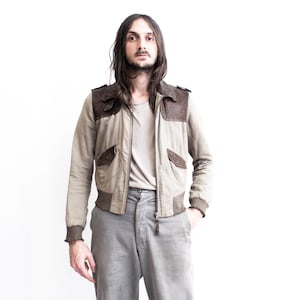 May include: A man wearing a beige and brown bomber jacket with a zipper closure and a grey t-shirt underneath. He is wearing grey pants and has long brown hair.