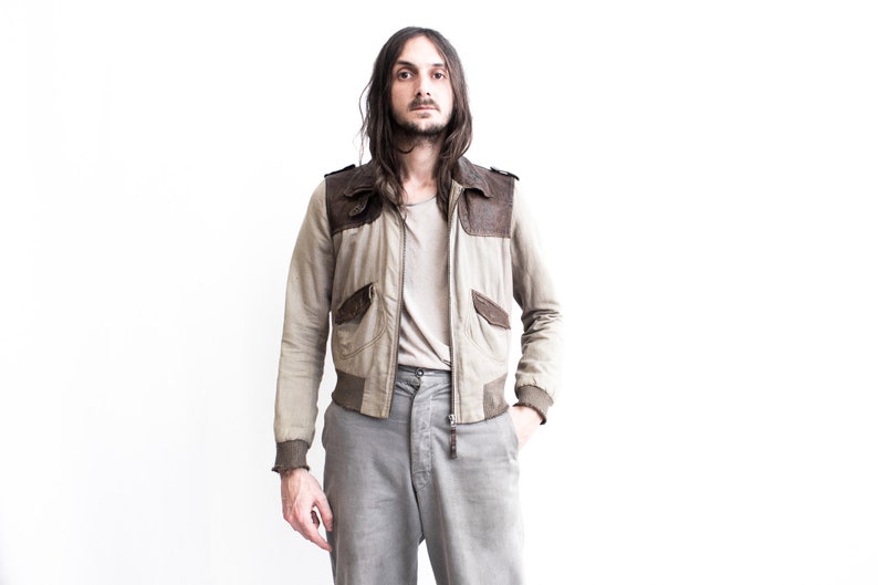 May include: A man wearing a beige and brown bomber jacket with a zipper closure and a grey t-shirt underneath. He is wearing grey pants and has long brown hair.