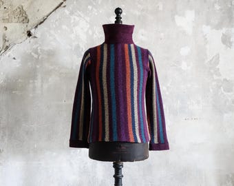 Puede incluir: Un jersey de cuello alto a rayas en tonos morado, verde azulado, naranja y beige. El jersey se exhibe en un maniquí sobre un fondo neutro y texturizado. El cuello alto y los puños son de color morado oscuro liso.