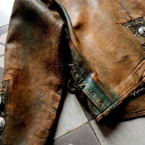 May include: A brown leather jacket with green trim and silver accents. The jacket has a zip fastening and a lace-up detail on the back.