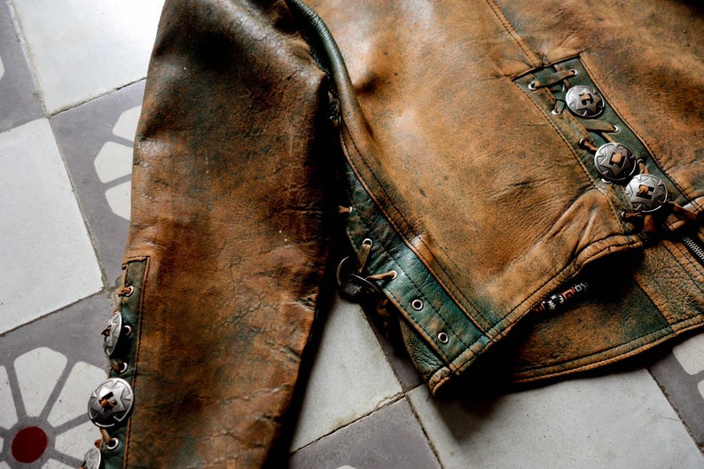 May include: A brown leather jacket with green trim and silver accents. The jacket has a zip fastening and a lace-up detail on the back.