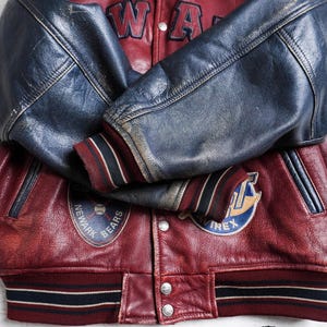 May include: A maroon and navy blue leather varsity jacket featuring long navy sleeves, striped ribbed cuffs, and a striped hem. The front displays patches including a Newark Bears baseball logo and Avirex branding, with metal snap buttons.