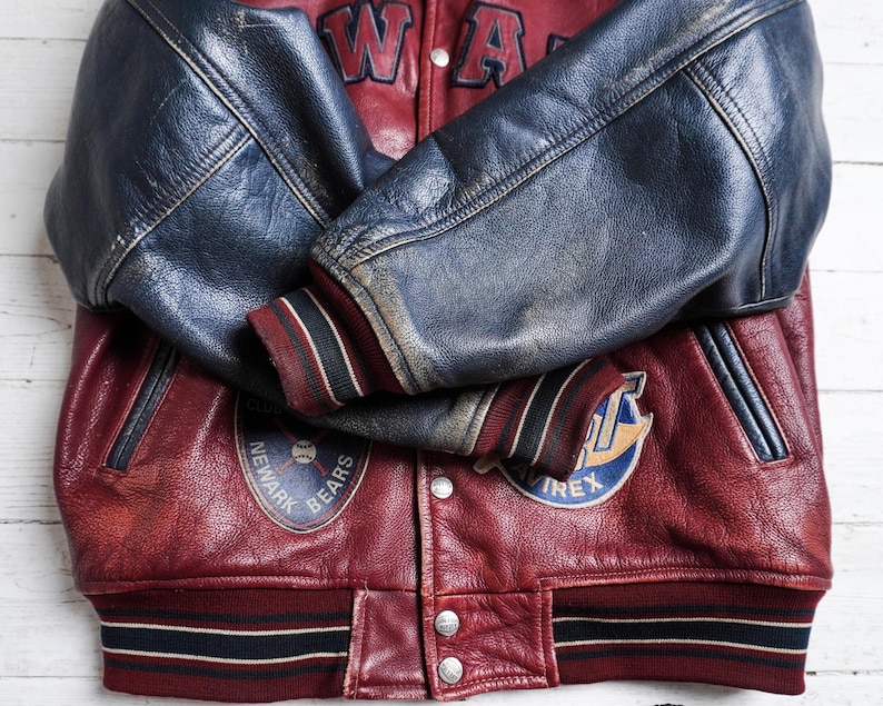 May include: A maroon and navy blue leather varsity jacket featuring long navy sleeves, striped ribbed cuffs, and a striped hem. The front displays patches including a Newark Bears baseball logo and Avirex branding, with metal snap buttons.