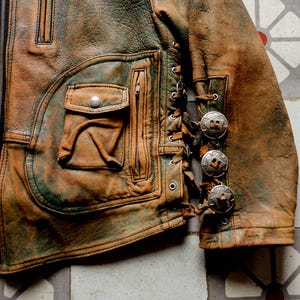 May include: A brown leather jacket with a zip fastening and a decorative lacing detail on the sleeve. The jacket has a large pocket on the front and several silver-toned metal accents.
