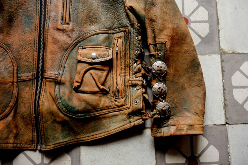 May include: A brown leather jacket with a zip fastening and a decorative lacing detail on the sleeve. The jacket has a large pocket on the front and several silver-toned metal accents.