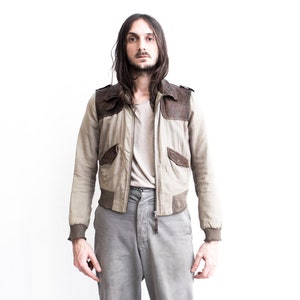 May include: A man wearing a light brown bomber jacket with brown leather accents and gray pants.