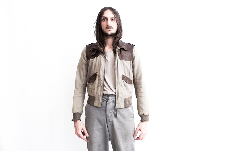 May include: A man wearing a light brown bomber jacket with brown leather accents and gray pants.