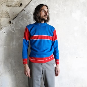 Könnte beinhalten: Ein blau-rotes Langarmshirt mit einem horizontalen roten Streifen auf der Brust. Das Shirt hat einen hohen Ausschnitt und weiße Verzierungen. Das Model trägt eine graue Hose.