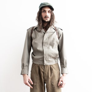Pu&ograve; includere: Un berretto e una giacca verde oliva vintage con pantaloni abbinati. La giacca ha un colletto, spalline e un cinturino in pelle marrone. I pantaloni sono di una tonalit&agrave; di marrone pi&ugrave; scura. L'outfit &egrave; indossato da una persona.