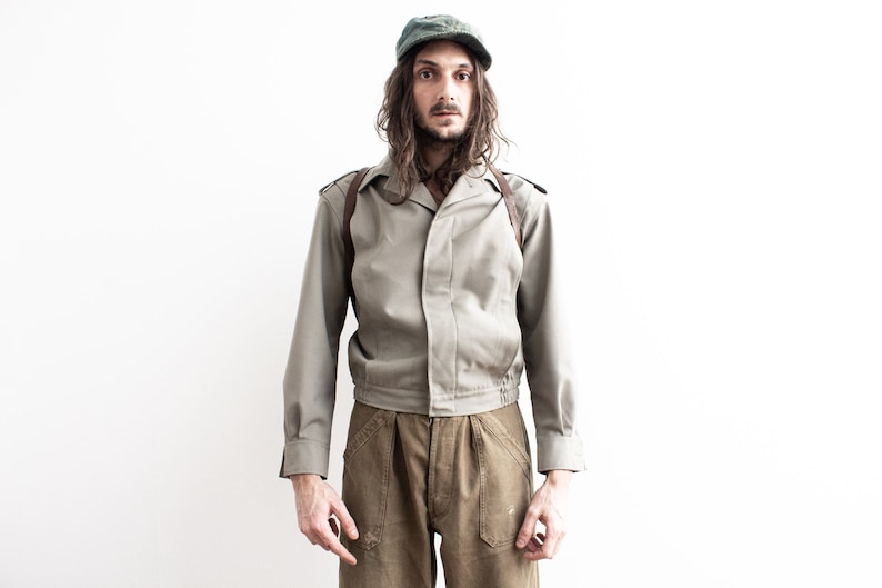 Pu&ograve; includere: Un berretto e una giacca verde oliva vintage con pantaloni abbinati. La giacca ha un colletto, spalline e un cinturino in pelle marrone. I pantaloni sono di una tonalit&agrave; di marrone pi&ugrave; scura. L'outfit &egrave; indossato da una persona.