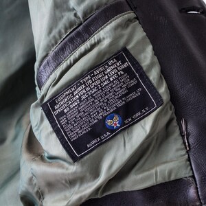 K&ouml;nnte beinhalten: Eine dunkelbraune Lederjacke mit hellgr&uuml;nem Innenfutter. Innen ist ein schwarzes, rechteckiges Etikett eingen&auml;ht, das den wei&szlig;en Text EQUIPMENT - TYPE - AVIREX USA, AUTHENTIC AVIREX LTD FLIGHT GARMENT und AVIREX U.S.A. NEW YORK, N.Y. tr&auml;gt.