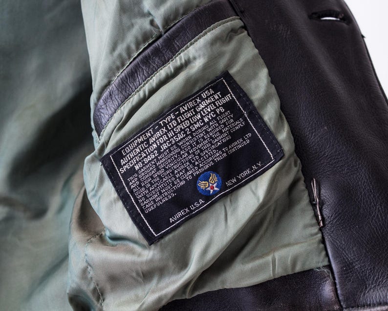 K&ouml;nnte beinhalten: Eine dunkelbraune Lederjacke mit hellgr&uuml;nem Innenfutter. Innen ist ein schwarzes, rechteckiges Etikett eingen&auml;ht, das den wei&szlig;en Text EQUIPMENT - TYPE - AVIREX USA, AUTHENTIC AVIREX LTD FLIGHT GARMENT und AVIREX U.S.A. NEW YORK, N.Y. tr&auml;gt.