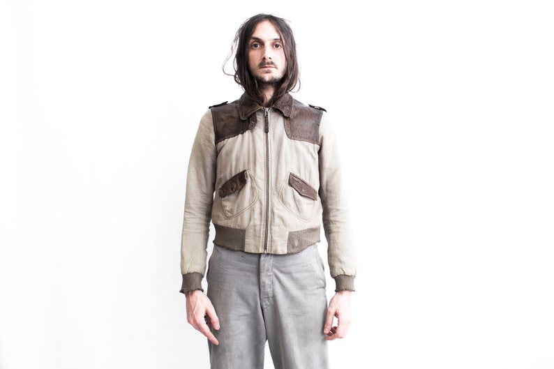 May include: A man wearing a light brown bomber jacket with brown leather accents and gray pants.