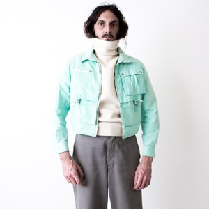 May include: A mint green cropped utility jacket featuring a front zipper closure, a pointed collar, and four large patch pockets with flaps. The jacket is worn over a cream-colored turtleneck sweater and paired with plain gray trousers.
