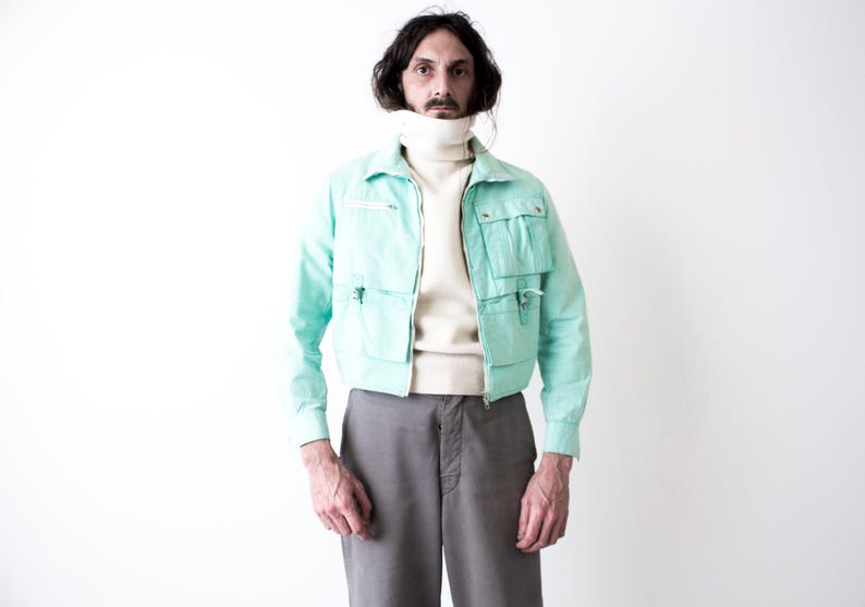 May include: A mint green cropped utility jacket featuring a front zipper closure, a pointed collar, and four large patch pockets with flaps. The jacket is worn over a cream-colored turtleneck sweater and paired with plain gray trousers.