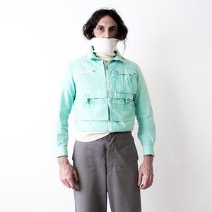 May include: A light turquoise jacket with multiple front pockets and a zipper closure. The jacket is worn over a white turtleneck and paired with gray pants. The overall style is vintage-inspired.