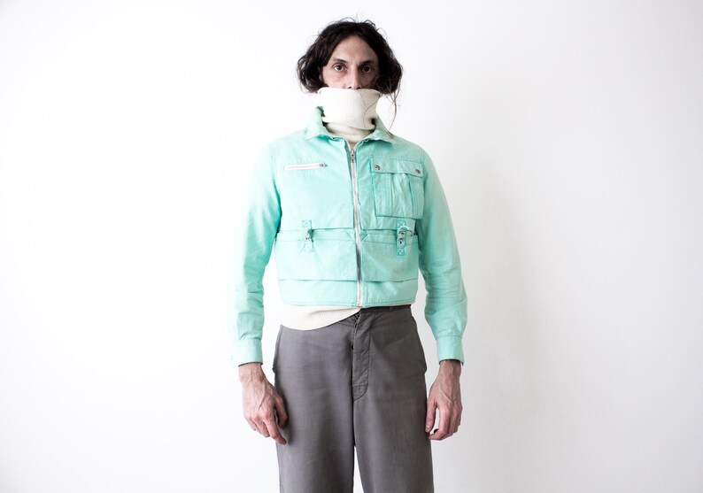 May include: A light turquoise jacket with multiple front pockets and a zipper closure. The jacket is worn over a white turtleneck and paired with gray pants. The overall style is vintage-inspired.