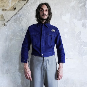 May include: A royal blue work jacket featuring a pointed collar, long sleeves with buttoned cuffs, and two chest pockets with flaps. An oval patch with the text SIP is visible on the upper left chest. The jacket has a buttoned front closure.
