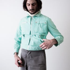 May include: A light turquoise jacket with multiple pockets and a zipper closure. The jacket has a collared neckline and long sleeves. The person is wearing gray pants and a white turtleneck. The jacket has a unique design.