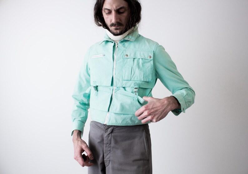May include: A light turquoise jacket with multiple pockets and a zipper closure. The jacket has a collared neckline and long sleeves. The person is wearing gray pants and a white turtleneck. The jacket has a unique design.