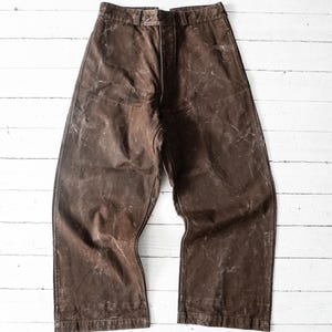 Può includere: Un paio di pantaloni marrone scuro caratterizzati da un design a vita alta con chiusura a bottone in vita. Il tessuto presenta un aspetto invecchiato e usurato con pieghe visibili e una finitura testurizzata su tutta la gamba larga.
