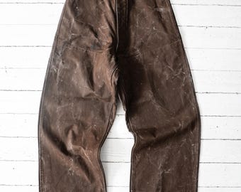 Può includere: Un paio di pantaloni marrone scuro caratterizzati da un design a vita alta con chiusura a bottone in vita. Il tessuto presenta un aspetto invecchiato e usurato con pieghe visibili e una finitura testurizzata su tutta la gamba larga.